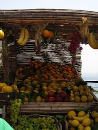 A market stall displays a variety of fresh fruits, including bananas, oranges, lemons, grapes, and tomatoes. The fruits are arranged in baskets made of woven bamboo, creating a rustic and inviting appearance. The fruits appear vibrant under the daylight, with leaves adding a natural touch.