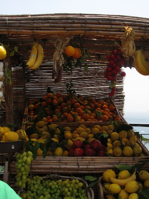 A market stall displays a variety of fresh fruits, including bananas, oranges, lemons, grapes, and tomatoes. The fruits are arranged in baskets made of woven bamboo, creating a rustic and inviting appearance. The fruits appear vibrant under the daylight, with leaves adding a natural touch.