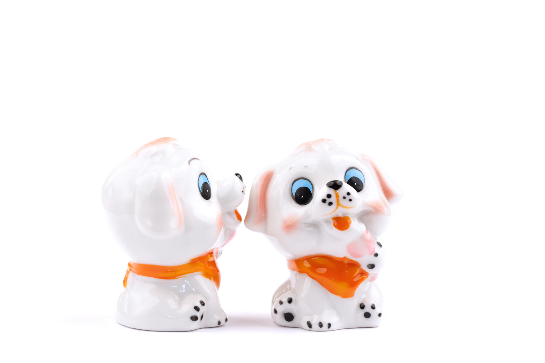 A pair of glossy white puppy figurines with blue eyes and orange scarves, nose to nose on a white surface