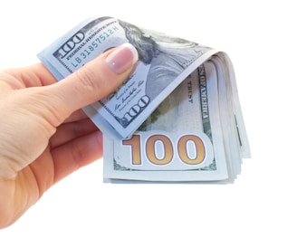A hand with well-manicured nails holds a stack of 100 dollar bills, with one bill prominently visible displaying the number 100.
