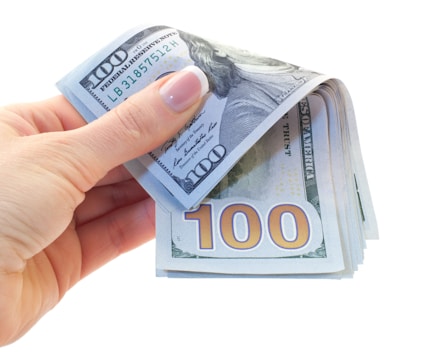 A hand with well-manicured nails holds a stack of 100 dollar bills, with one bill prominently visible displaying the number 100.