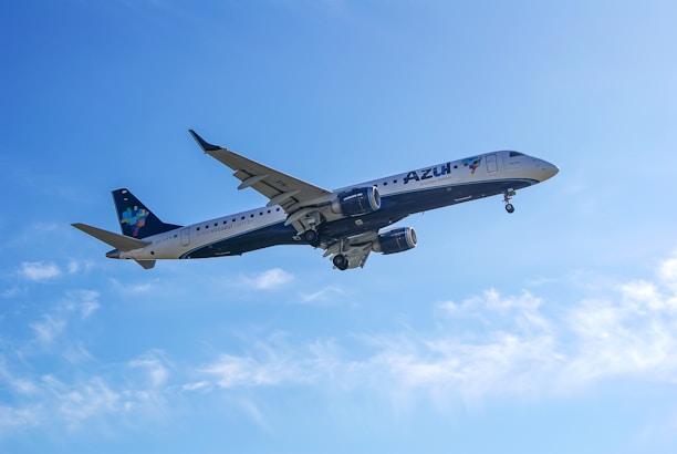 A commercial airplane is flying through a clear blue sky. The aircraft has visible engines under the wings and its tail is marked with colorful branding. The sunlight illuminates the plane's exterior, highlighting the logo.