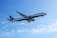 A commercial airplane is flying through a clear blue sky. The aircraft has visible engines under the wings and its tail is marked with colorful branding. The sunlight illuminates the plane's exterior, highlighting the logo.