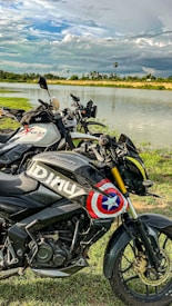 Three motorcycles are parked on a grassy area by a serene lake, with a cloudy sky in the background. The motorcycle in the foreground has a custom design with a colorful graphic that resembles a shield.
