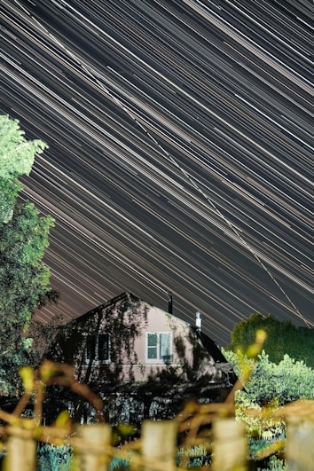 A long exposure photograph capturing the streaks of stars moving across the night sky above a house surrounded by trees. The house is partially obscured by shadows and foliage, creating a serene and dynamic celestial scene.