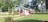 A small, single-story house with a white and pink exterior is surrounded by neatly trimmed grass and a variety of plants. The house features a sloped roof and is situated amidst a lush, green landscape with several large trees in the background.