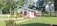 A small, single-story house with a white and pink exterior is surrounded by neatly trimmed grass and a variety of plants. The house features a sloped roof and is situated amidst a lush, green landscape with several large trees in the background.