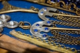 A collection of ornate metallic objects, including items with blue gemstones and intricate chain designs, are displayed on a blue fabric surface. The pieces appear to be embellished with detailed craftsmanship.