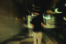A person is walking down a dimly lit, narrow alley at night. The image is blurred, giving it a sense of motion and surrealism. There are various streaks of light and reflections along the walls, emphasizing the dark and mysterious atmosphere.