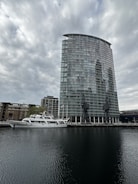 A modern, glass high-rise building stands near the edge of a body of water, with two large cranes positioned in front. A white yacht is docked alongside, with several older brick buildings visible in the background. The sky is overcast, casting a subdued light over the scene.