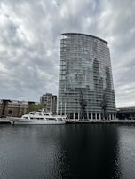 A modern, glass high-rise building stands near the edge of a body of water, with two large cranes positioned in front. A white yacht is docked alongside, with several older brick buildings visible in the background. The sky is overcast, casting a subdued light over the scene.