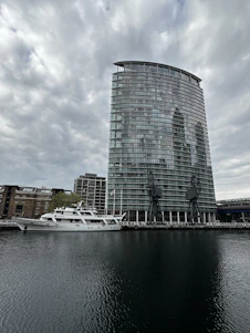 A modern, glass high-rise building stands near the edge of a body of water, with two large cranes positioned in front. A white yacht is docked alongside, with several older brick buildings visible in the background. The sky is overcast, casting a subdued light over the scene.