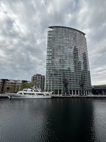 A modern, glass high-rise building stands near the edge of a body of water, with two large cranes positioned in front. A white yacht is docked alongside, with several older brick buildings visible in the background. The sky is overcast, casting a subdued light over the scene.