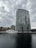 A modern, glass high-rise building stands near the edge of a body of water, with two large cranes positioned in front. A white yacht is docked alongside, with several older brick buildings visible in the background. The sky is overcast, casting a subdued light over the scene.