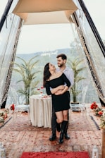 A couple embraces joyfully in a romantic setting inside a tent decorated with string lights and flower petals. A small table, set with wine glasses and scattered petals, sits behind them. Two palm plants flank the couple, adding greenery to the scene. A proposal message, 'Will you marry me?' is visible above them.