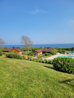 A scenic landscape with a manicured lawn leading to several wooden cabins surrounded by lush greenery. The ocean is visible in the background under a clear blue sky. There are some trees and a swimming pool with lounge chairs nearby.