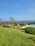 A scenic landscape with a manicured lawn leading to several wooden cabins surrounded by lush greenery. The ocean is visible in the background under a clear blue sky. There are some trees and a swimming pool with lounge chairs nearby.