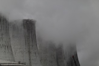 A group of large industrial cooling towers partially obscured by dense fog and steam. The structures appear weathered, with dark streaks running down their surfaces, indicating exposure to the elements. The sky is overcast, contributing to a grey and gloomy atmosphere.