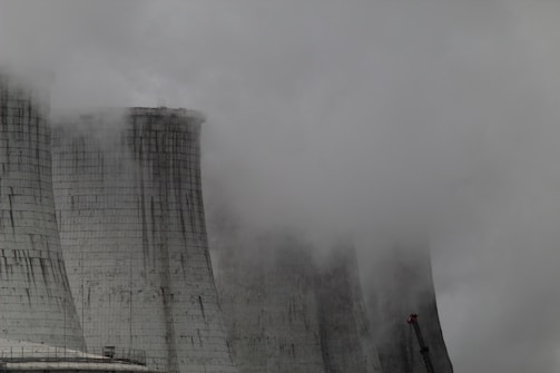 A group of large industrial cooling towers partially obscured by dense fog and steam. The structures appear weathered, with dark streaks running down their surfaces, indicating exposure to the elements. The sky is overcast, contributing to a grey and gloomy atmosphere.