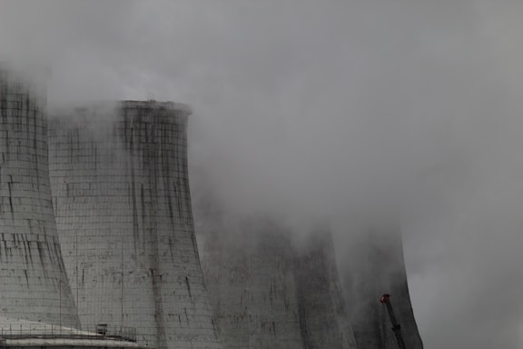 A group of large industrial cooling towers partially obscured by dense fog and steam. The structures appear weathered, with dark streaks running down their surfaces, indicating exposure to the elements. The sky is overcast, contributing to a grey and gloomy atmosphere.