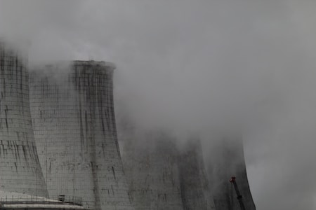 A group of large industrial cooling towers partially obscured by dense fog and steam. The structures appear weathered, with dark streaks running down their surfaces, indicating exposure to the elements. The sky is overcast, contributing to a grey and gloomy atmosphere.