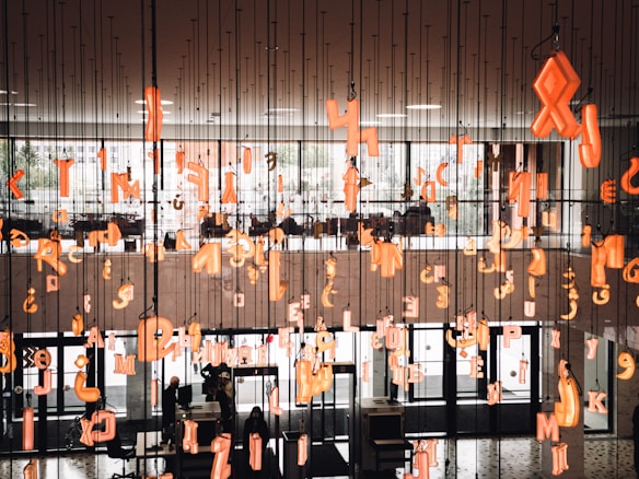 An indoor installation with numerous suspended orange neon alphabet letters and symbols hanging from the ceiling. In the background, there is a large window letting in natural light, and below, people are moving around a lobby area. The environment has a modern architectural style with glass doors and black interiors.