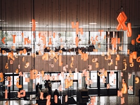 An indoor installation with numerous suspended orange neon alphabet letters and symbols hanging from the ceiling. In the background, there is a large window letting in natural light, and below, people are moving around a lobby area. The environment has a modern architectural style with glass doors and black interiors.