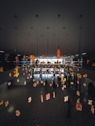 The scene features a modern interior space with a mirrored ceiling and floor reflecting various illuminated letters suspended in the air. People are walking beneath this installation, creating an artistic and immersive environment.