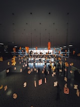 The scene features a modern interior space with a mirrored ceiling and floor reflecting various illuminated letters suspended in the air. People are walking beneath this installation, creating an artistic and immersive environment.
