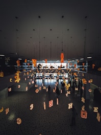 The scene features a modern interior space with a mirrored ceiling and floor reflecting various illuminated letters suspended in the air. People are walking beneath this installation, creating an artistic and immersive environment.