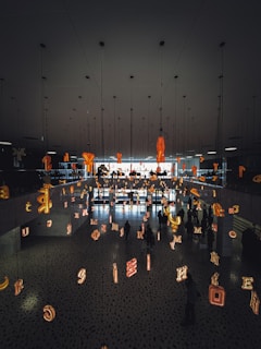 The scene features a modern interior space with a mirrored ceiling and floor reflecting various illuminated letters suspended in the air. People are walking beneath this installation, creating an artistic and immersive environment.
