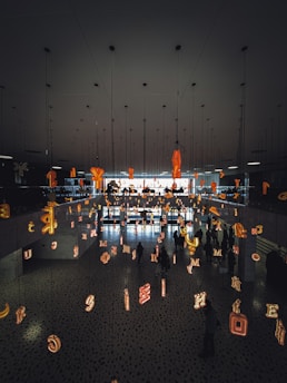 The scene features a modern interior space with a mirrored ceiling and floor reflecting various illuminated letters suspended in the air. People are walking beneath this installation, creating an artistic and immersive environment.