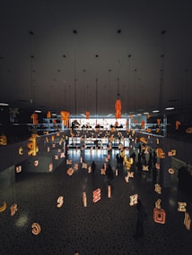 The scene features a modern interior space with a mirrored ceiling and floor reflecting various illuminated letters suspended in the air. People are walking beneath this installation, creating an artistic and immersive environment.