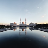 A large white mosque with multiple domes and minarets stands majestically, reflecting perfectly in a still pool of water in the foreground. The architecture is intricate and symmetrical, set against a clear sky transitioning from blue to a hint of orange, suggesting early morning or late evening.