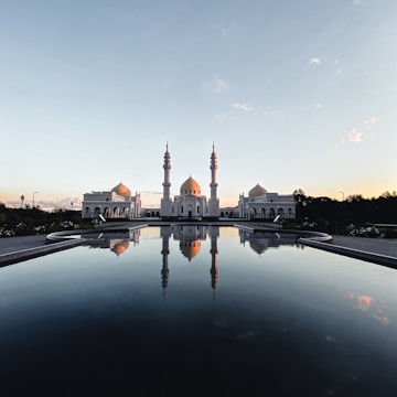 A large white mosque with multiple domes and minarets stands majestically, reflecting perfectly in a still pool of water in the foreground. The architecture is intricate and symmetrical, set against a clear sky transitioning from blue to a hint of orange, suggesting early morning or late evening.