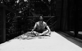 A person is sitting cross-legged in a meditative pose on a sunlit deck. The surroundings include a railing with wires and some trees casting shadows. The scene has a tranquil and serene ambiance.