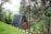 A small, wooden A-frame cabin nestled amidst lush greenery and tall trees. The cabin is surrounded by dense forestation, with a sloping grassy area in the foreground. The setting appears tranquil and secluded, suggesting a peaceful retreat.