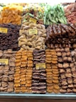 An assortment of dried fruits and nuts is neatly displayed. There are varieties such as dried apricots, figs, muska, and tropical fruits, all carefully arranged in rows with clear labels indicating their types. The colors range from bright oranges to deep browns and greens, creating a vibrant display.