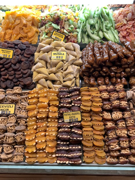 An assortment of dried fruits and nuts is neatly displayed. There are varieties such as dried apricots, figs, muska, and tropical fruits, all carefully arranged in rows with clear labels indicating their types. The colors range from bright oranges to deep browns and greens, creating a vibrant display.