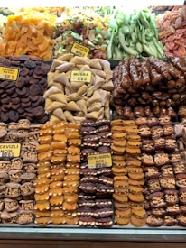 An assortment of dried fruits and nuts is neatly displayed. There are varieties such as dried apricots, figs, muska, and tropical fruits, all carefully arranged in rows with clear labels indicating their types. The colors range from bright oranges to deep browns and greens, creating a vibrant display.