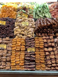 An assortment of dried fruits and nuts is neatly displayed. There are varieties such as dried apricots, figs, muska, and tropical fruits, all carefully arranged in rows with clear labels indicating their types. The colors range from bright oranges to deep browns and greens, creating a vibrant display.