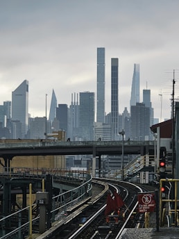 A cityscape featuring a view of tall skyscrapers in the distance under a cloudy sky. In the foreground, there is an elevated railway track with a red warning flag and a sign that reads 'Do not enter or cross tracks.' The area is surrounded by urban infrastructure, including bridges and railing.