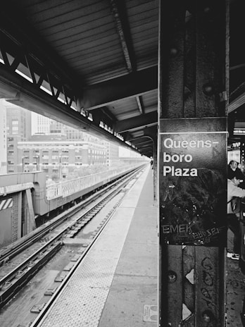 A black and white photograph of an elevated train platform with tracks running parallel to the platform. In the foreground, there is a metal pillar with graffiti and the words 'Queensboro Plaza' visible. The platform is mostly empty, with a few people partially visible on the right side. Buildings can be seen in the background beyond the platform.