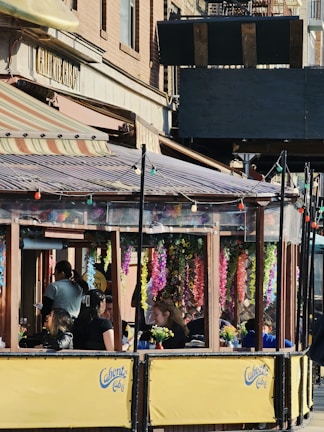 An outdoor cafe with a wooden canopy adorned with vibrant, hanging flower garlands. Several people are seated at tables, engaged in conversation or looking at menus. The yellow barriers have 'Caliente Cab Co' branding, and colorful string lights are strung across. The cafe is located along a brick-lined street.