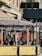 An outdoor cafe with a wooden canopy adorned with vibrant, hanging flower garlands. Several people are seated at tables, engaged in conversation or looking at menus. The yellow barriers have 'Caliente Cab Co' branding, and colorful string lights are strung across. The cafe is located along a brick-lined street.