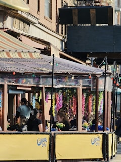 An outdoor cafe with a wooden canopy adorned with vibrant, hanging flower garlands. Several people are seated at tables, engaged in conversation or looking at menus. The yellow barriers have 'Caliente Cab Co' branding, and colorful string lights are strung across. The cafe is located along a brick-lined street.