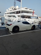 A shiny luxury sports car parked by a marina with yachts in the background under a golden sunset.