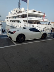 A luxury sports car parked near a large yacht, with both the car and yacht displaying sleek, modern designs. The setting appears to be a marina with calm water and other boats visible in the background.