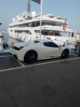A luxury sports car parked near a large yacht, with both the car and yacht displaying sleek, modern designs. The setting appears to be a marina with calm water and other boats visible in the background.