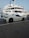 A luxury sports car parked near a large yacht, with both the car and yacht displaying sleek, modern designs. The setting appears to be a marina with calm water and other boats visible in the background.